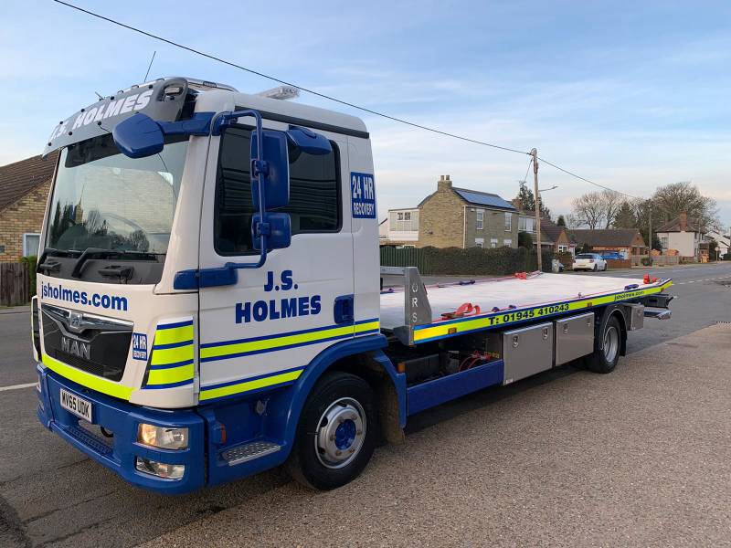 J.S. Holmes recovery truck parked on a road. The truck is white with blue and red stripes, and it has the words "J.S. HOLMES" and "24 HR RECOVERY" 
