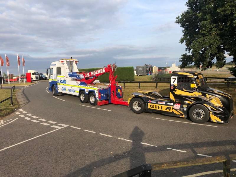 J.S. Holmes recovery truck towing a racing truck on a road. The racing truck is black and yellow with the number 7 on the side. 